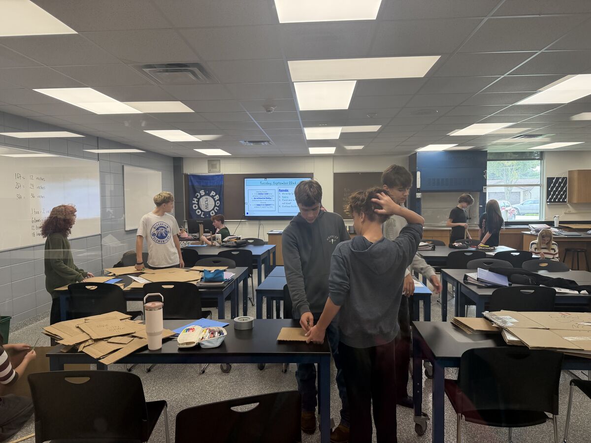 Students work together on a group project in a bright classroom, cutting and shaping cardboard.