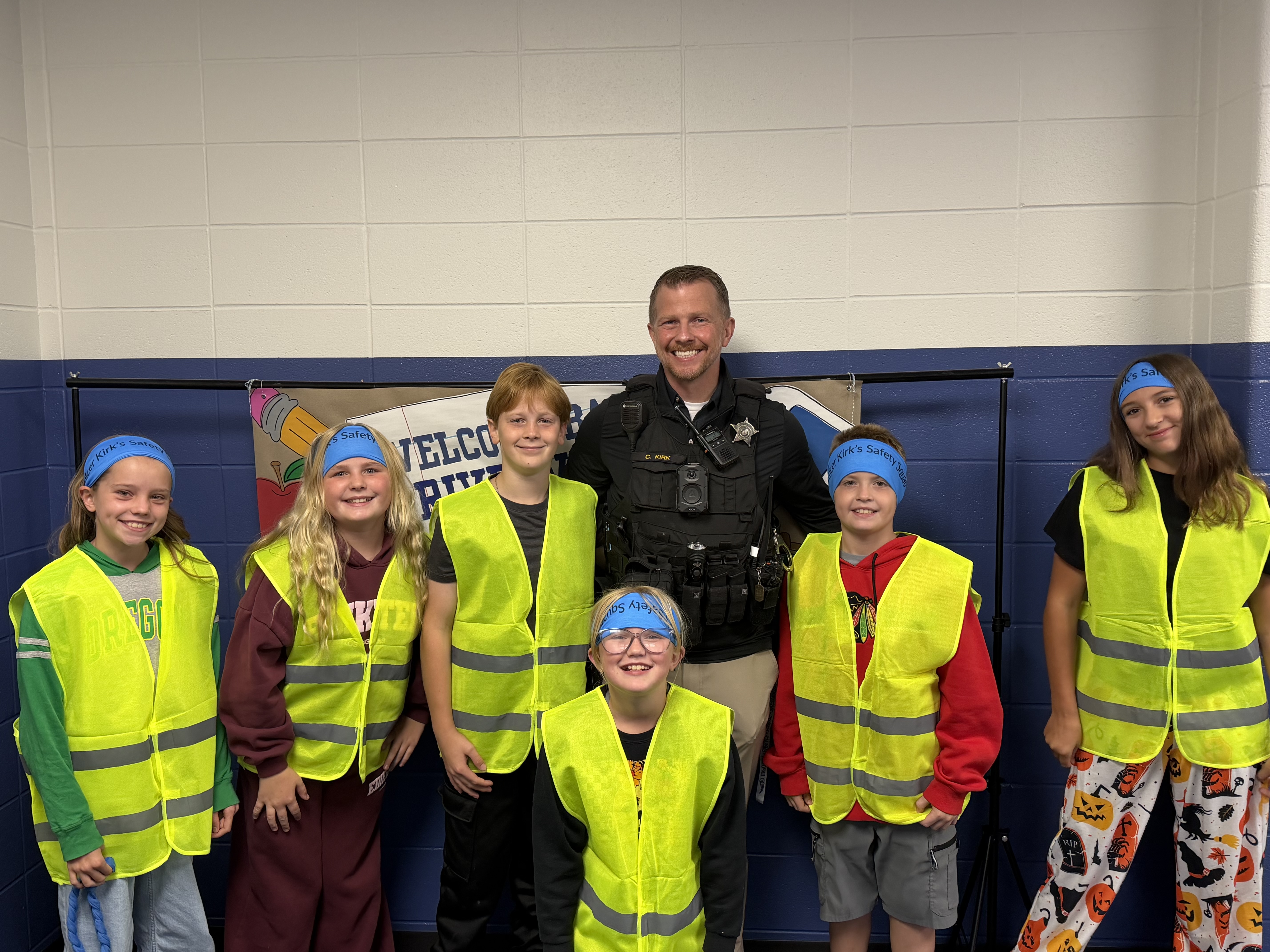 A smiling school safety officer stands with six children wearing neon safety vests and blue headbands in a hallway.