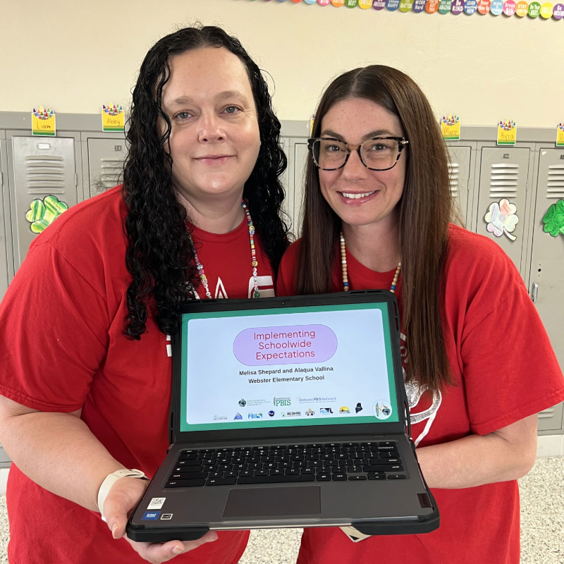 Two women in red shirts hold a laptop displaying "Implementing Schoolwide Expectations" at Webster Elementary School.