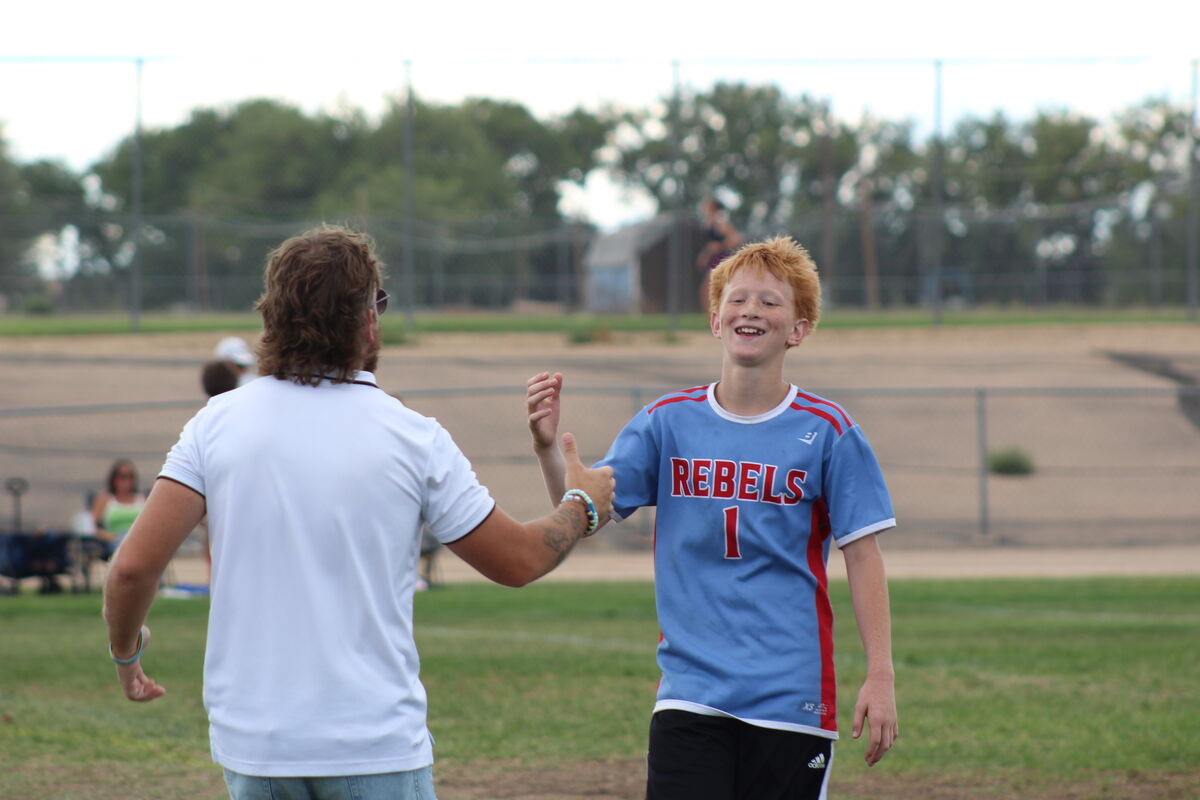A young boy in a blue and red "REBELS 1" jersey smiles as he gives a high-five to a coach in a white polo shirt on a grassy field.