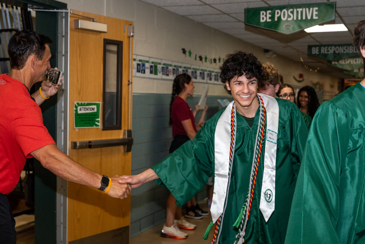A smiling graduate in a green cap and gown shakes hands with a man in a red shirt.