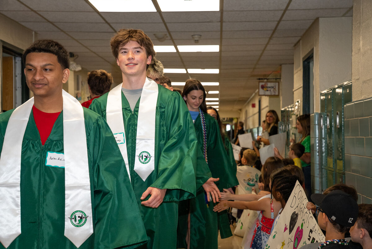 Graduates in green gowns and white sashes walk down a school hallway, greeted by cheering onlookers.