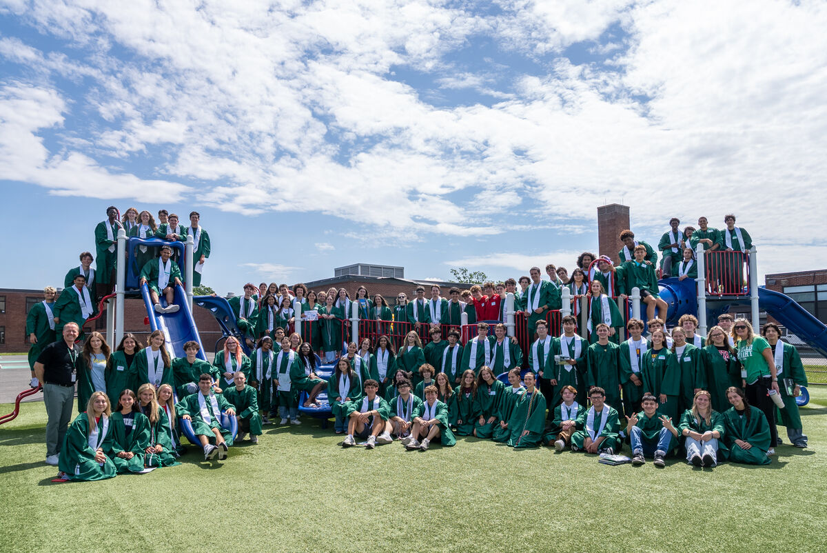 A large group of graduates in green gowns and white sashes pose for a photo on a playground.