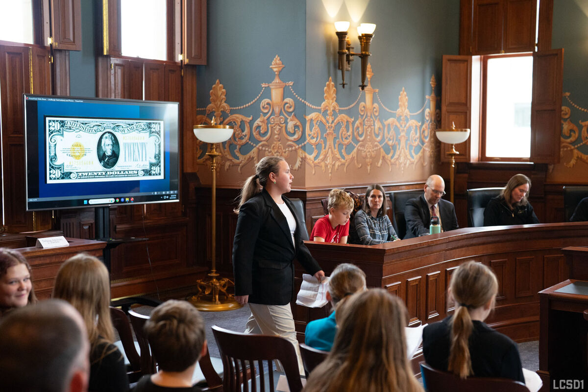 A student in a suit stands in front of a TV displaying a dollar bill.