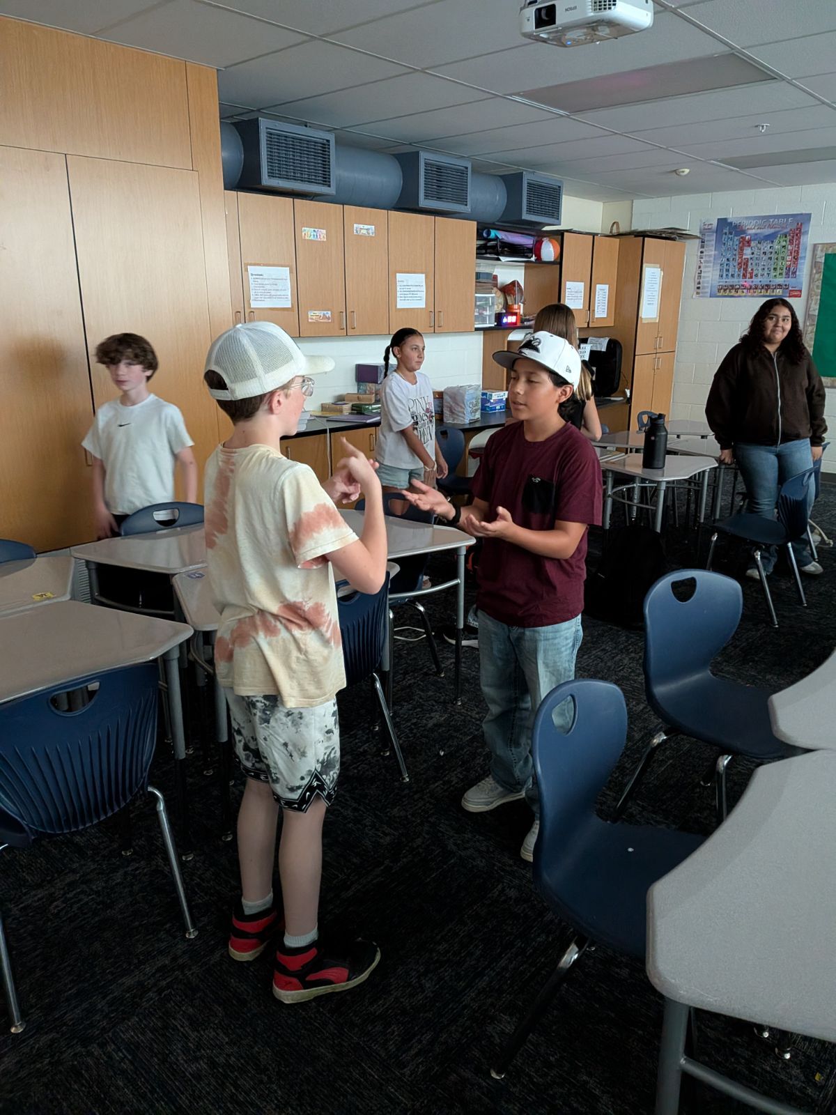 Two boys converse using sign language in a classroom while other students stand nearby.