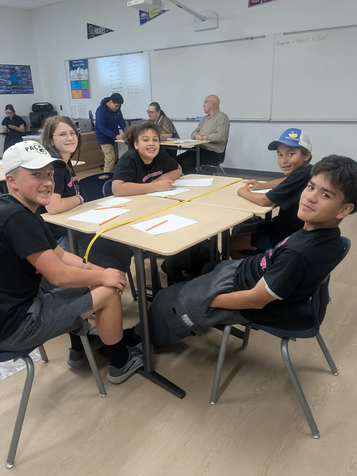 Five students sit at connected desks in a classroom, smiling and looking toward the camera.
