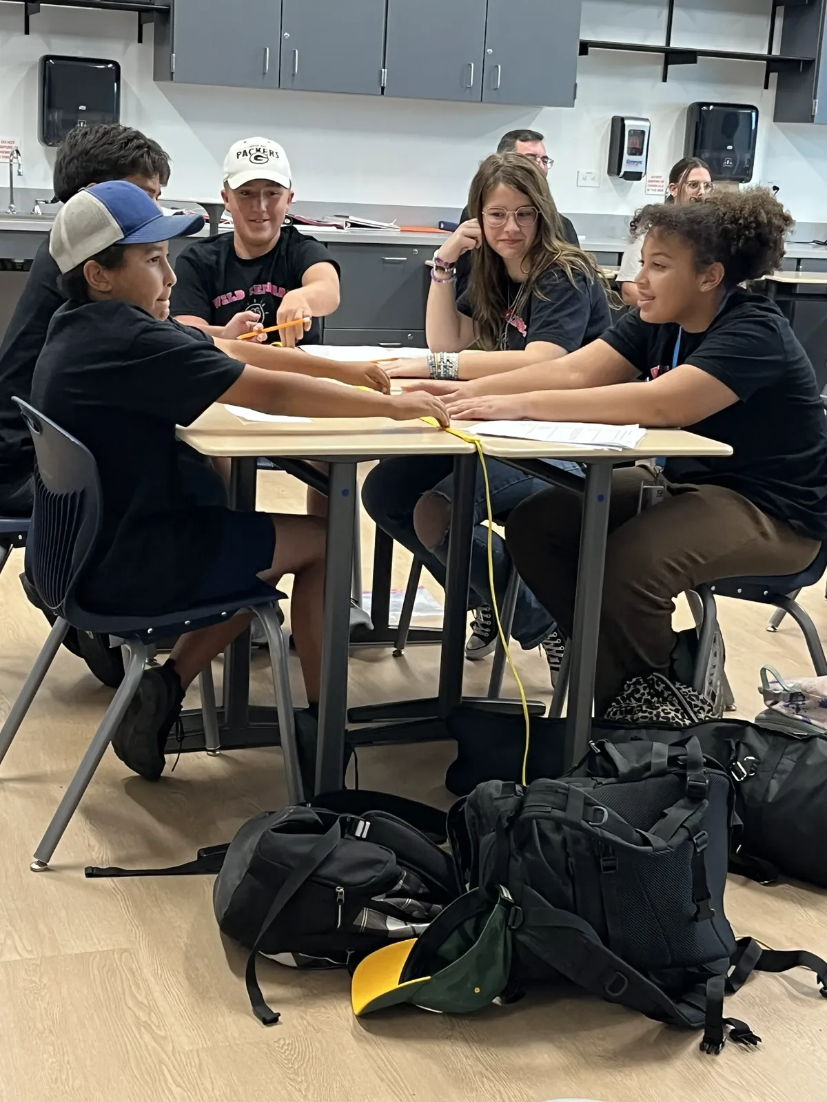 Students collaborate at a table in a classroom, stretching a yellow string together.