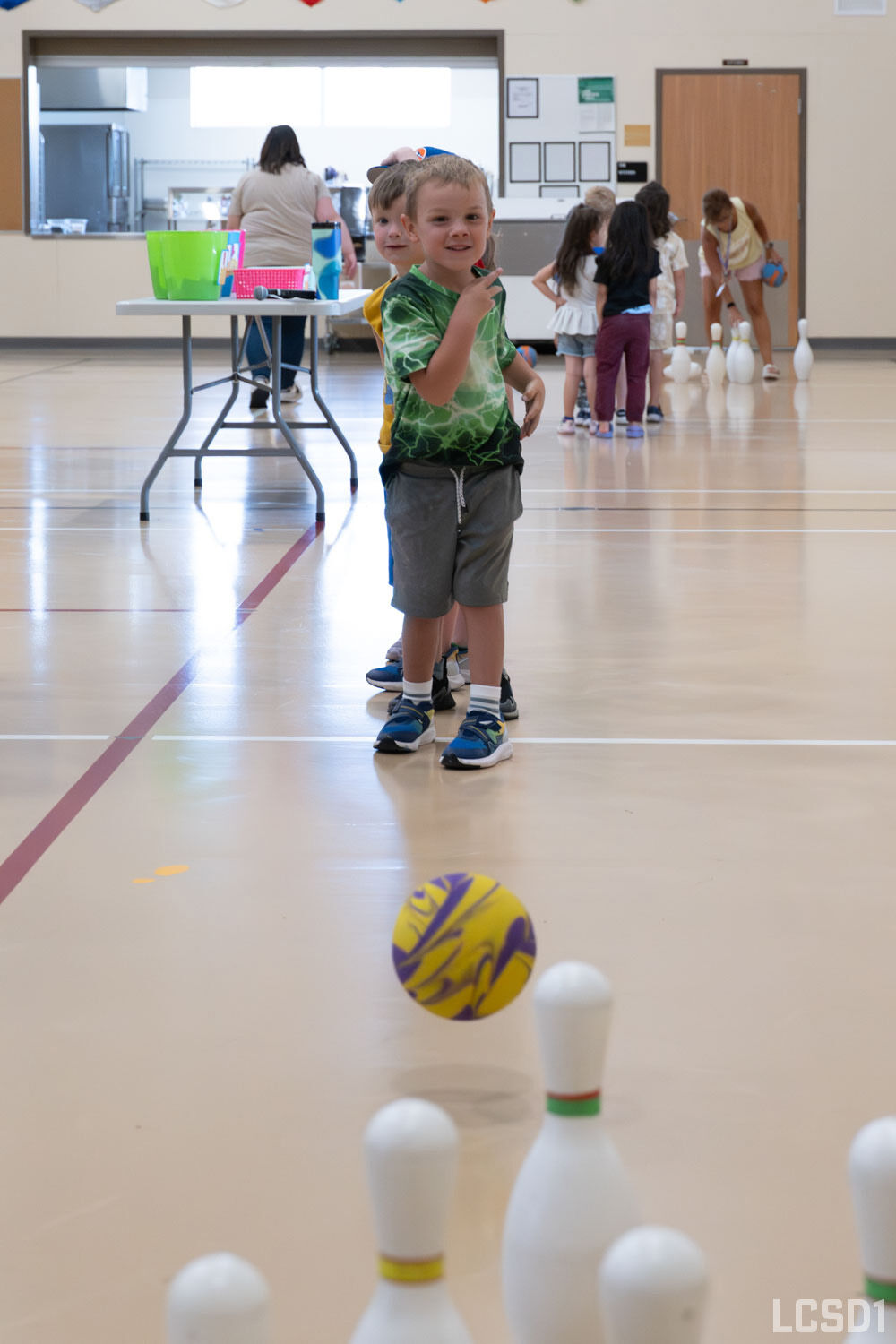 Children line up in a gymnasium to play a game of bowling with a colorful ball and white pins.