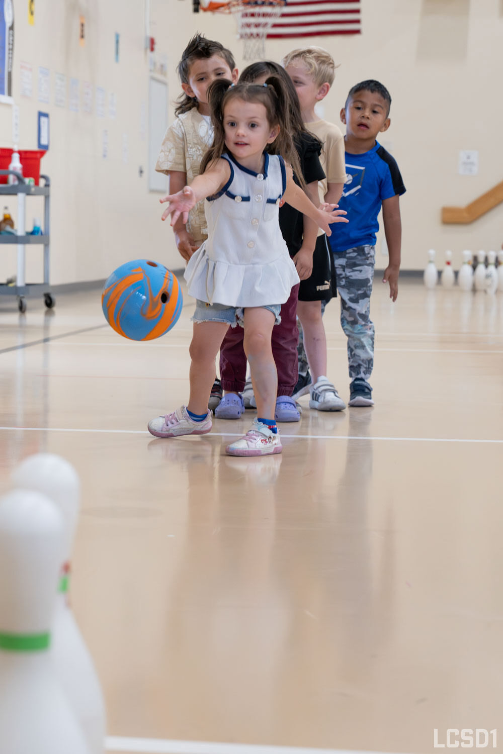 A young girl throws a bowling ball while other children watch in a gymnasium.