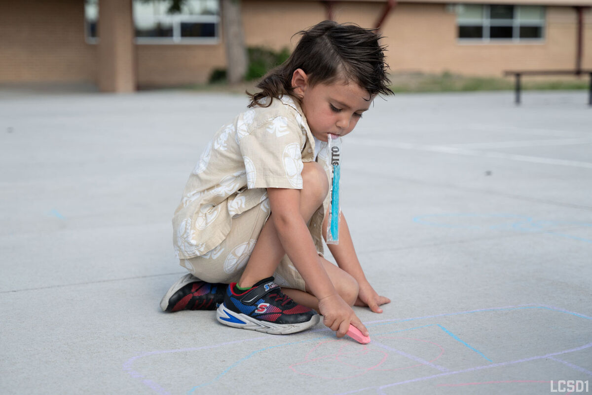 A young child kneels on a paved surface, drawing with pink chalk.