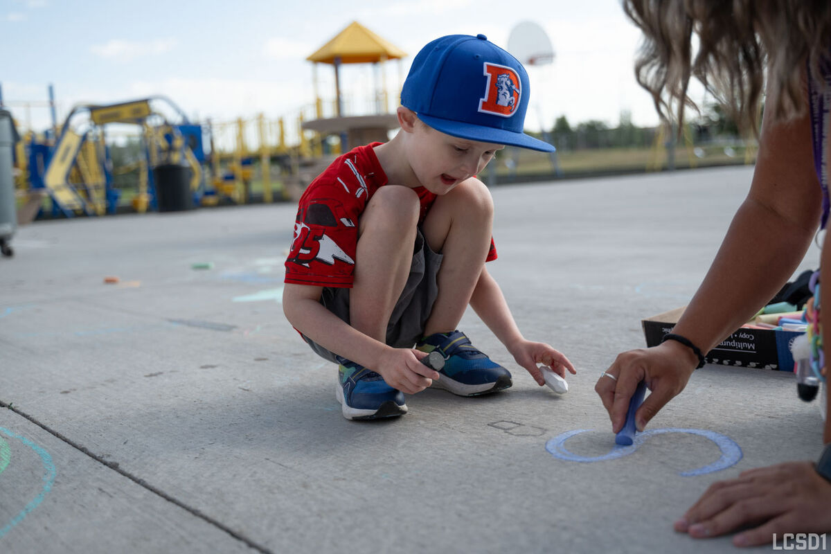A young boy in a blue baseball cap draws with chalk on a paved surface.