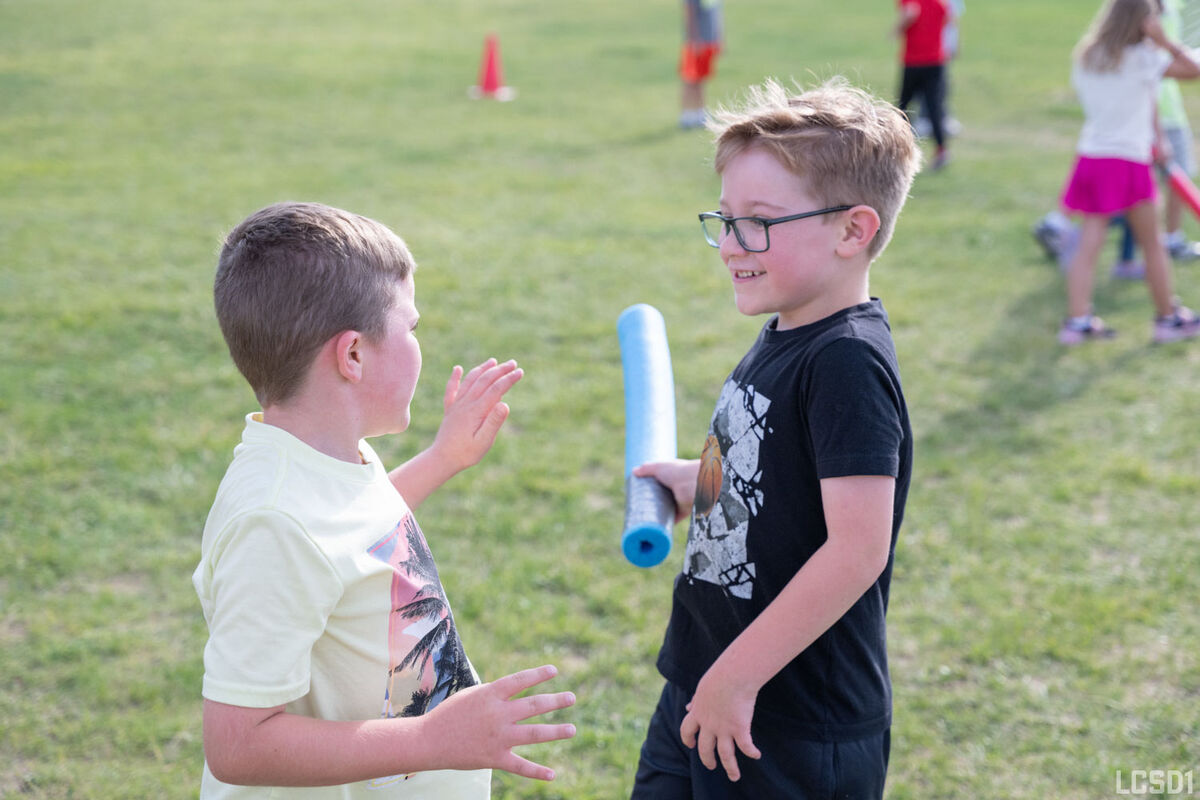 Two young boys interact on a grassy field, one holding a blue foam roller.
