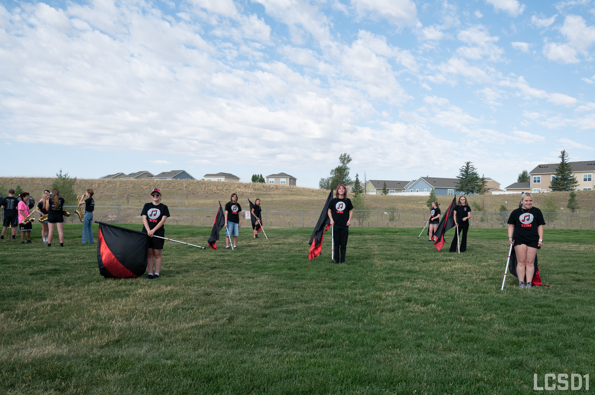 A group of young people in black t-shirts with a musical note logo practice with flags and saxophones on a grassy field.