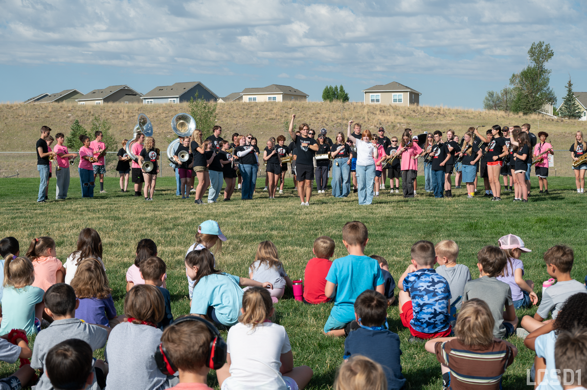 A marching band performs outdoors on a grassy field with an audience of children seated in front.