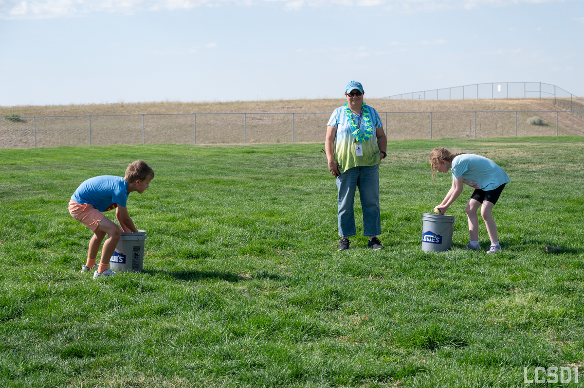 A woman in a tie-dye shirt and lei watches two children play a game with buckets on a grassy field.