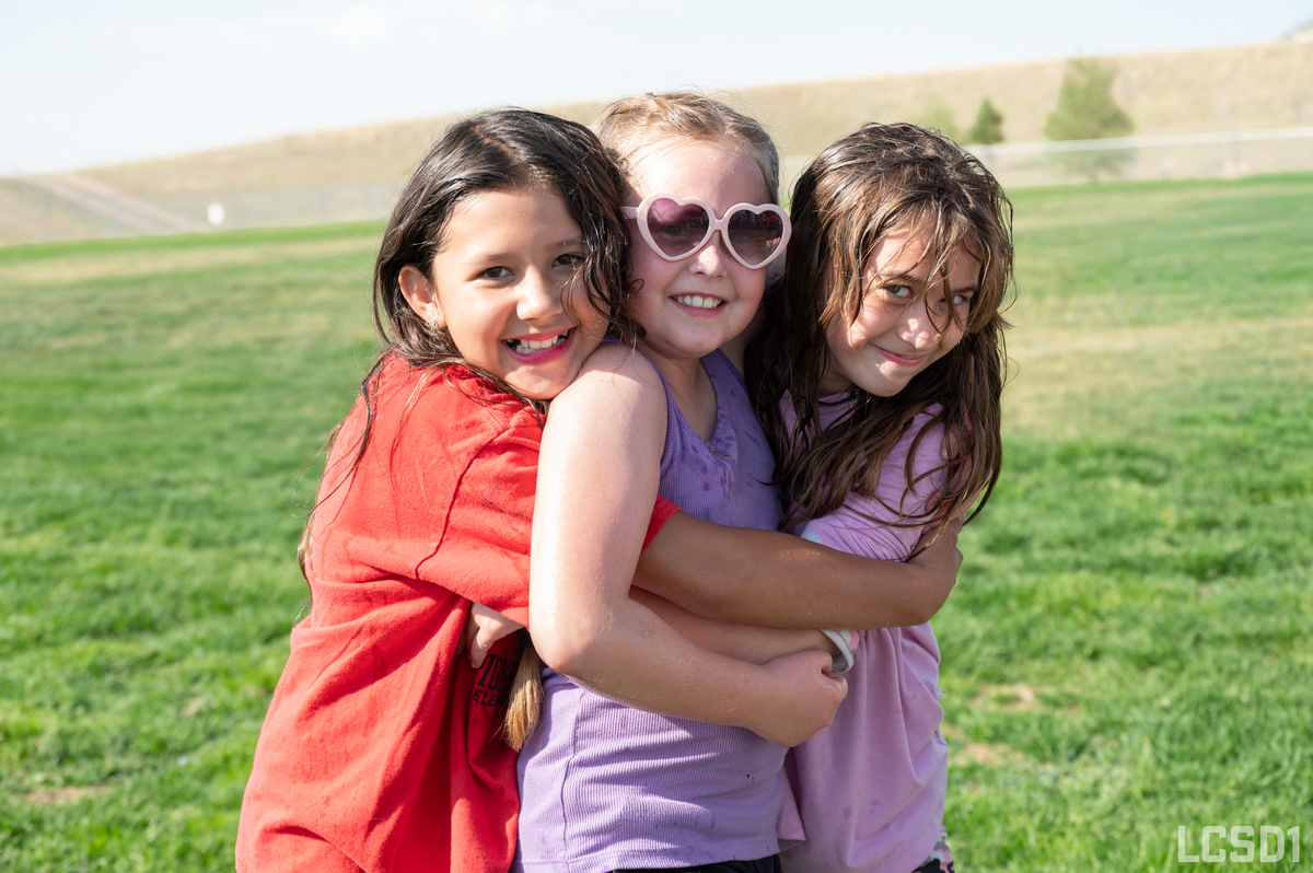 Three young girls in colorful shirts hug each other tightly outdoors on a grassy field.