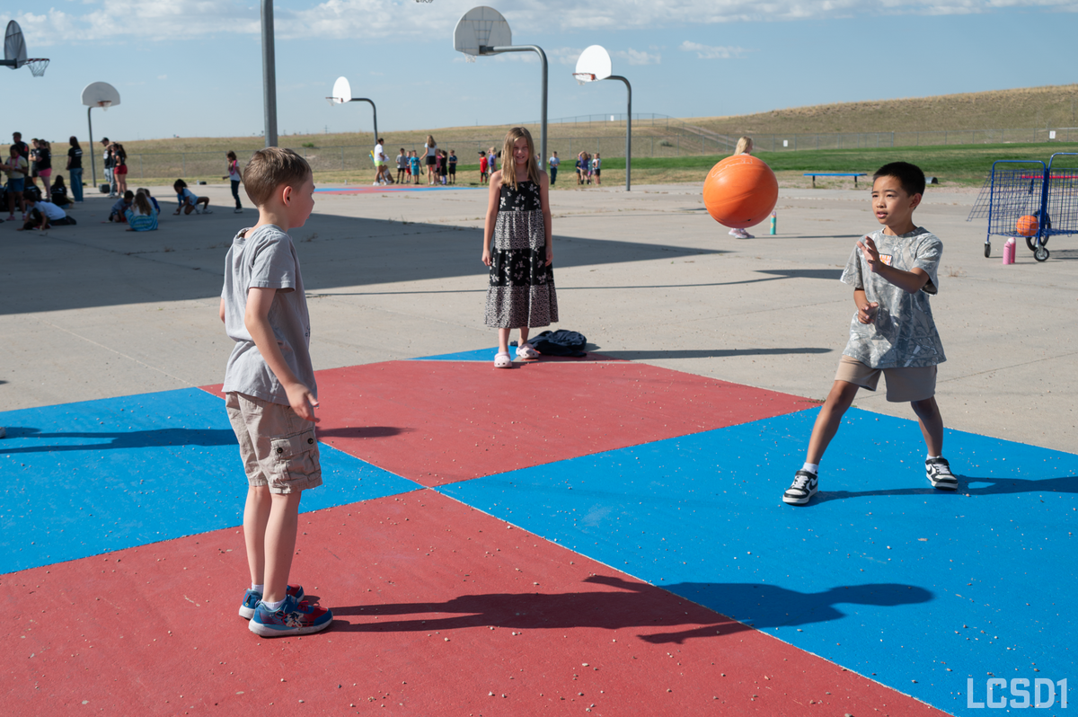 Two children play with an orange ball on a colorful outdoor court with basketball hoops.