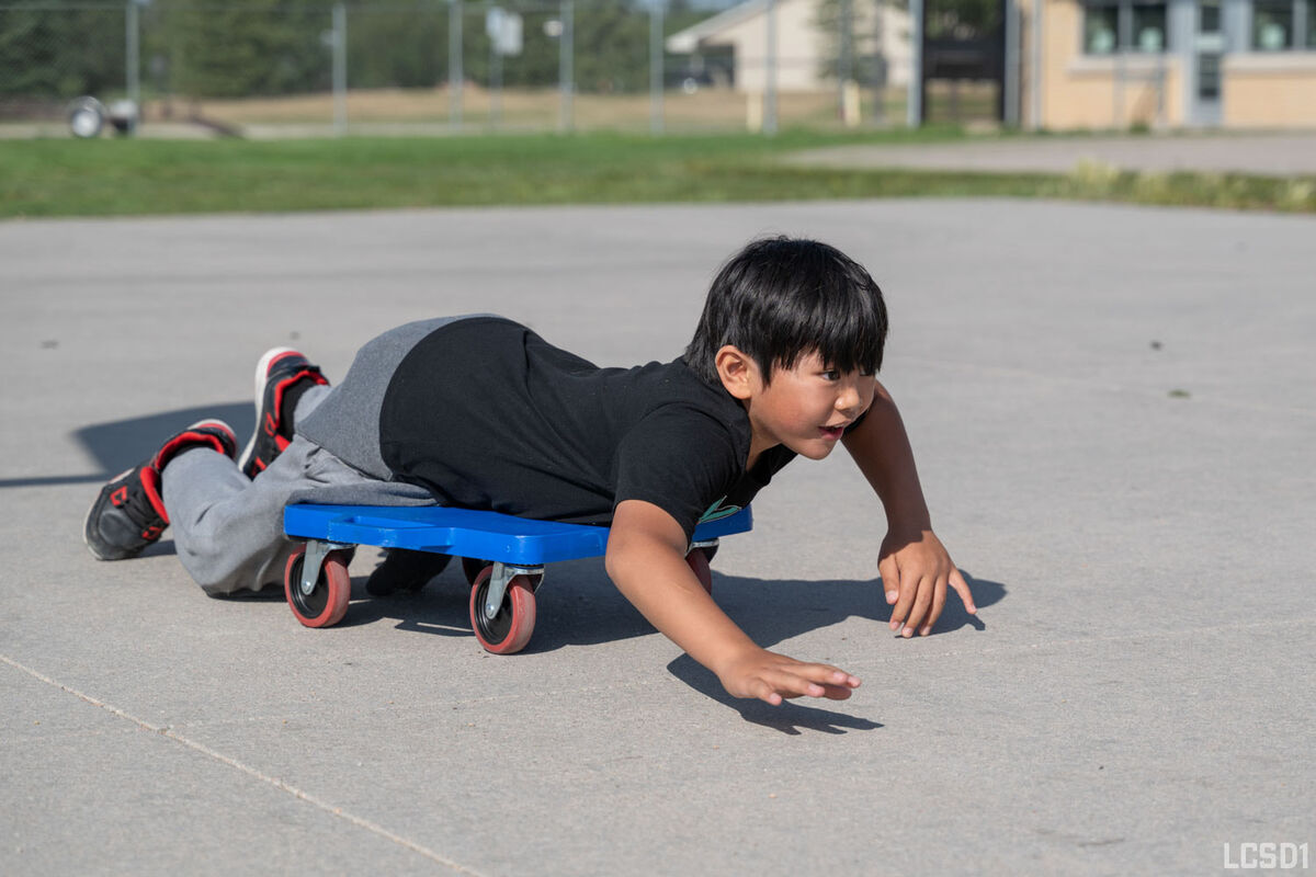 A young boy lies on a blue rolling scooter, propelling himself forward with his hands on a paved surface.