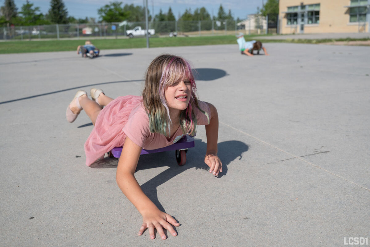 A young girl with colorful streaks in her hair lies on a purple scooter on a paved surface.