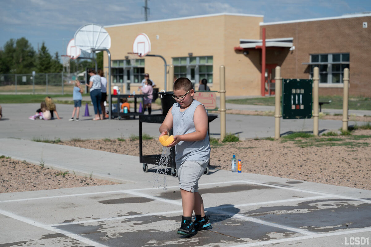 A young boy in a gray tank top and shorts squeezes a yellow sponge, splashing water on a paved schoolyard.