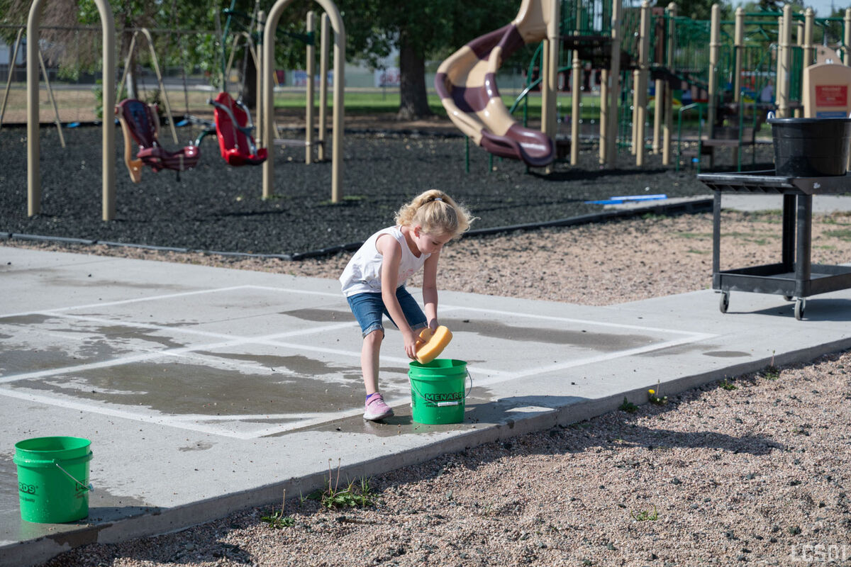 A young girl dips a sponge into a green bucket on a sunny day at a playground.