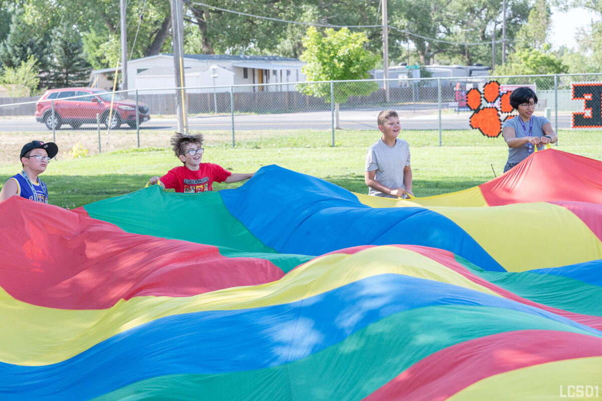 Children and an adult hold the edges of a large, colorful parachute on a grassy field.