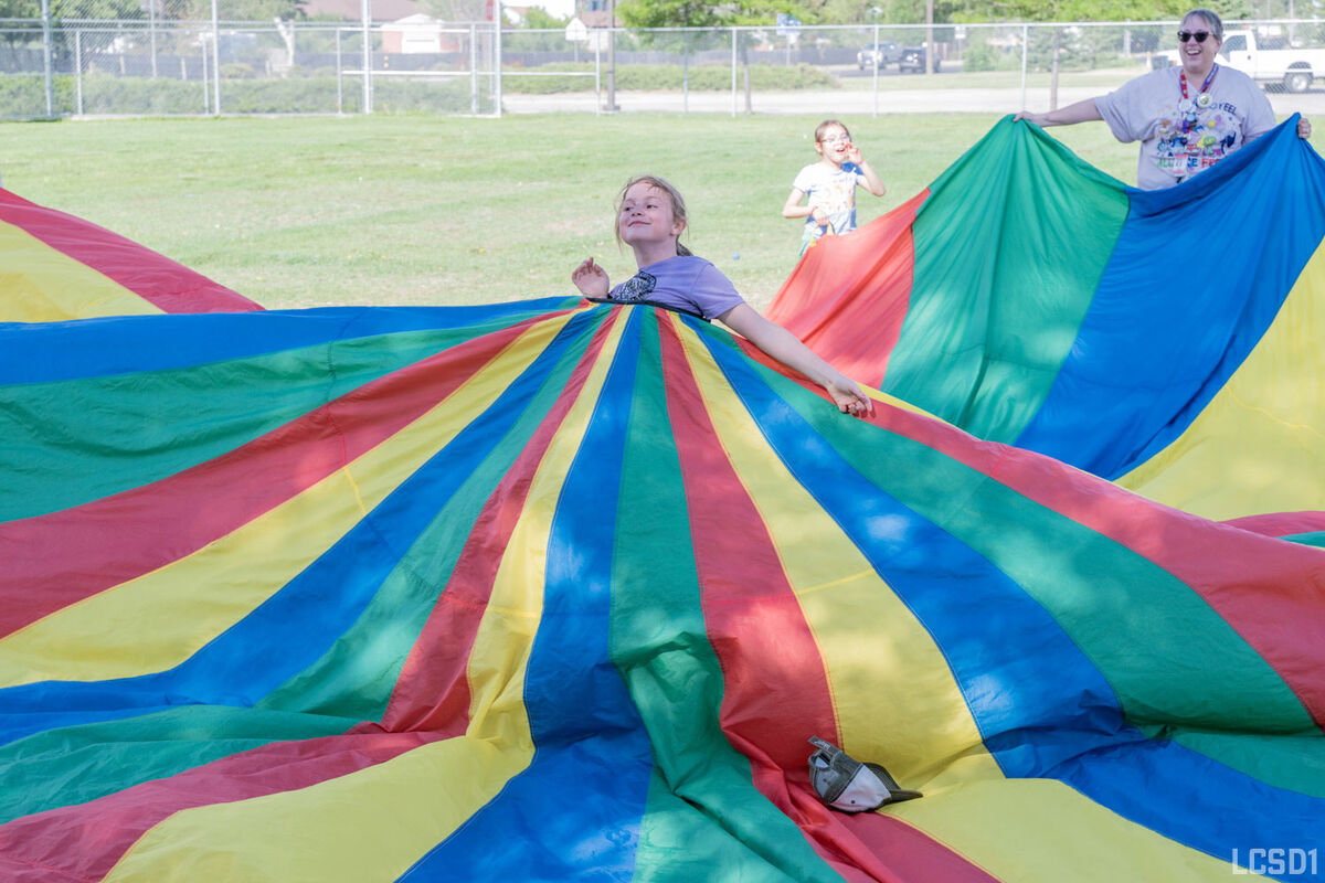 Children and adults play together under a large, colorful parachute on a grassy field.