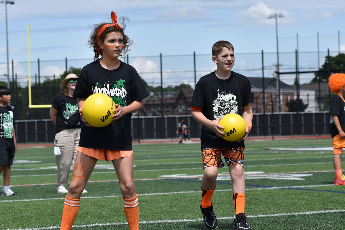 Two children stand on a green turf field, each holding a yellow ball, with a football goal in the background.