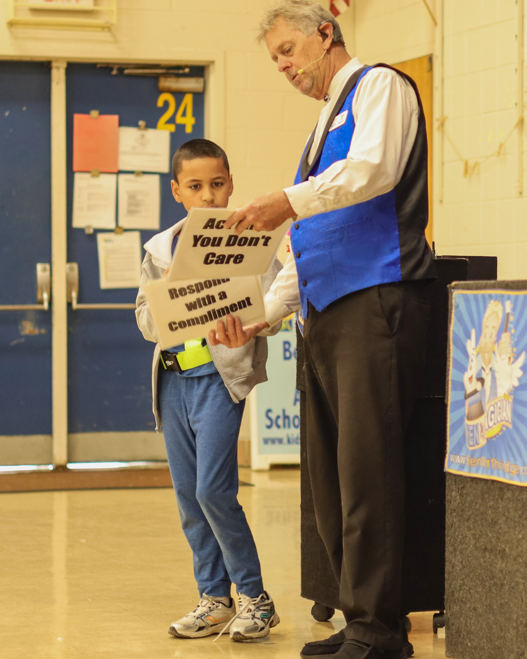 A young boy and an adult presenter hold up signs with text during an indoor school assembly.