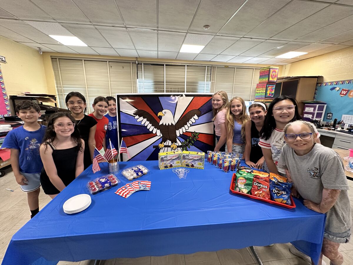 A group of smiling children stand around a table laden with snacks and a patriotic eagle banner.