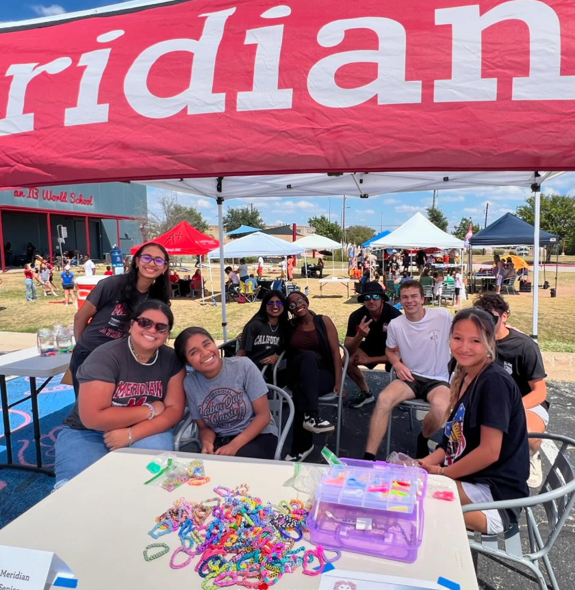 A group of smiling students sits around a table covered in colorful rubber band bracelets under a large red Meridian canopy.