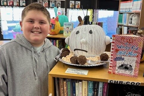 A smiling boy stands beside a creative pumpkin decorated as the character from "Diary of a Wimpy Kid: Hot Mess" in a library.