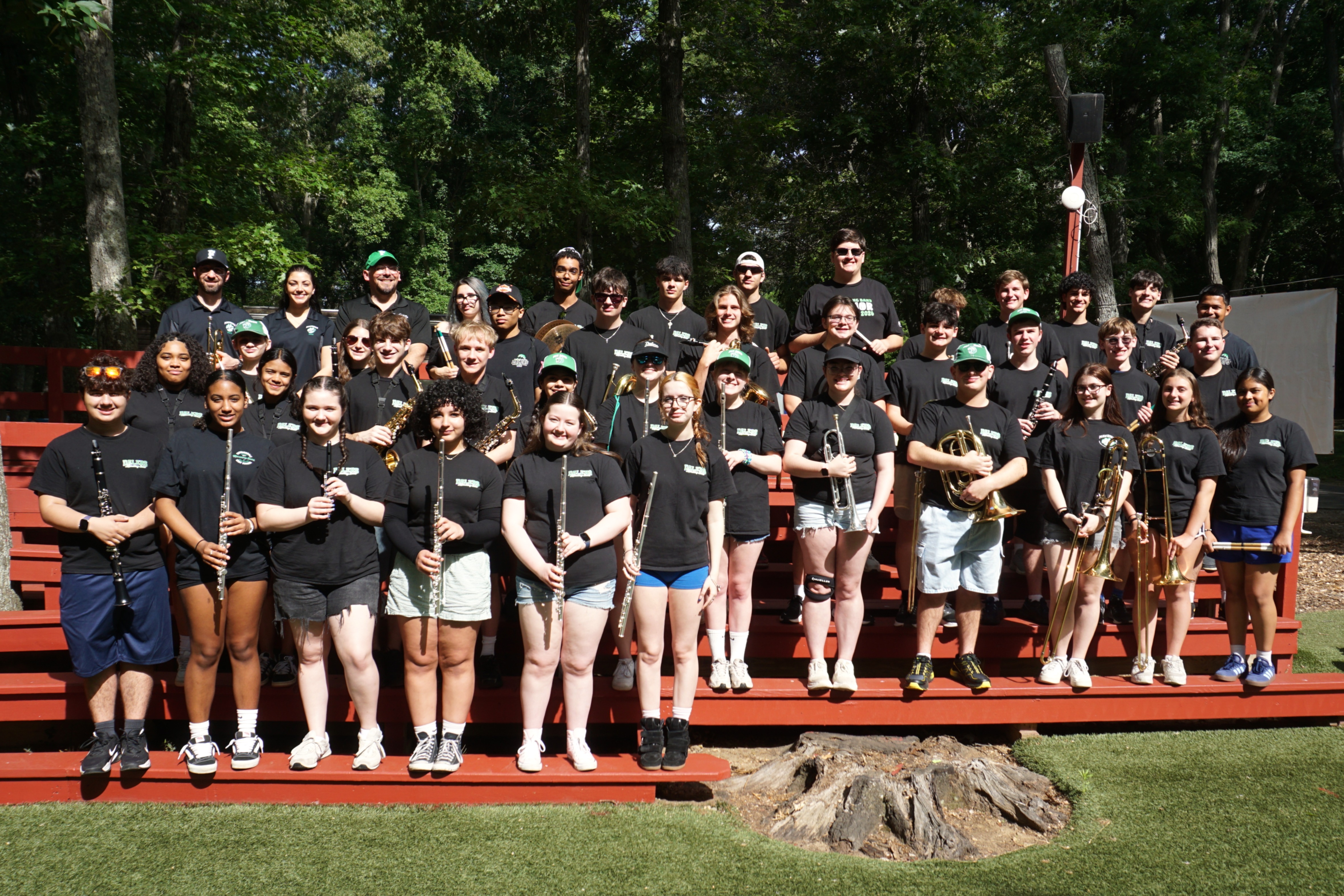 A diverse group of young musicians in black t-shirts and shorts pose for a photo outdoors, holding various brass and woodwind instruments.