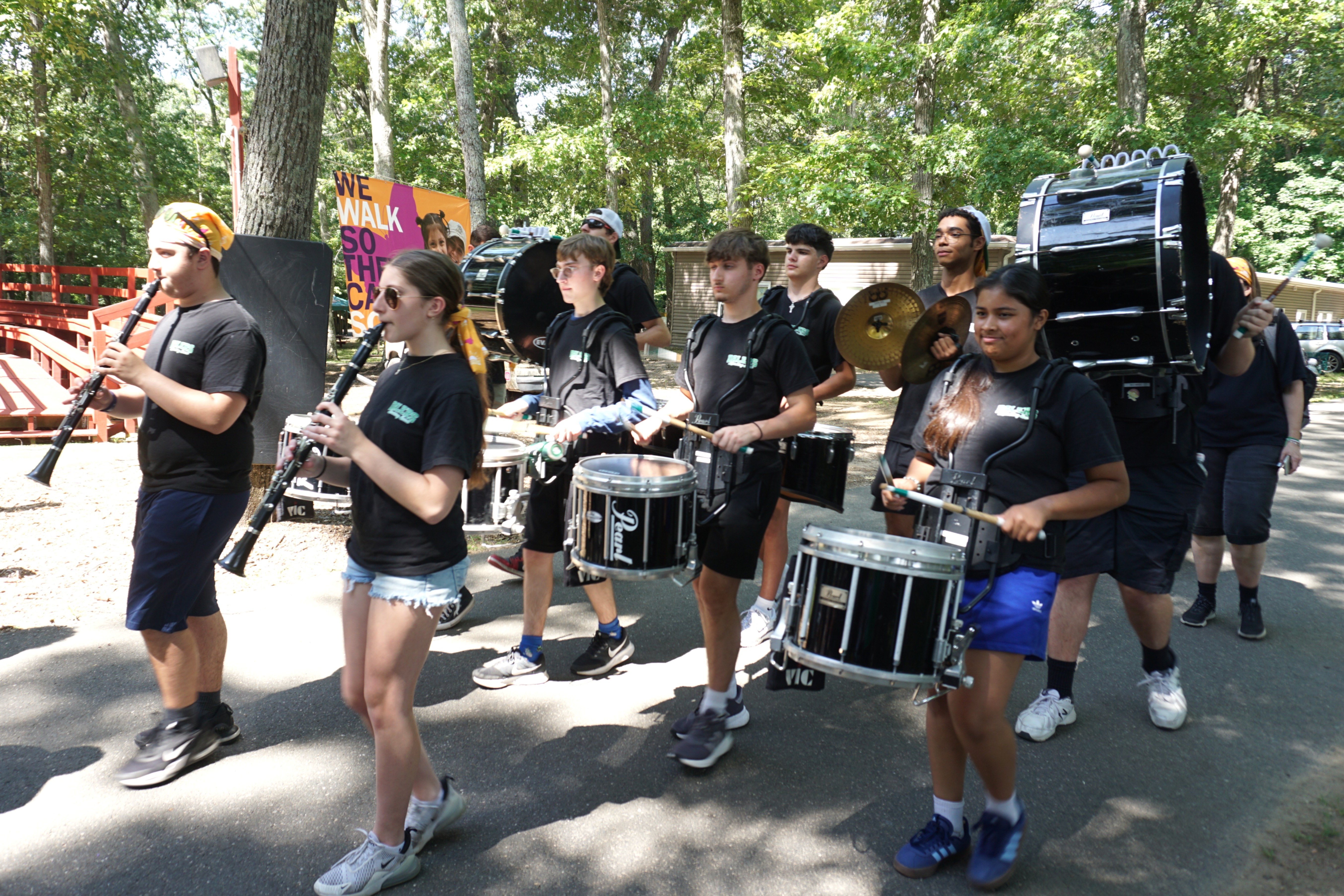 A marching band with drums and clarinets walks outdoors on a paved path.