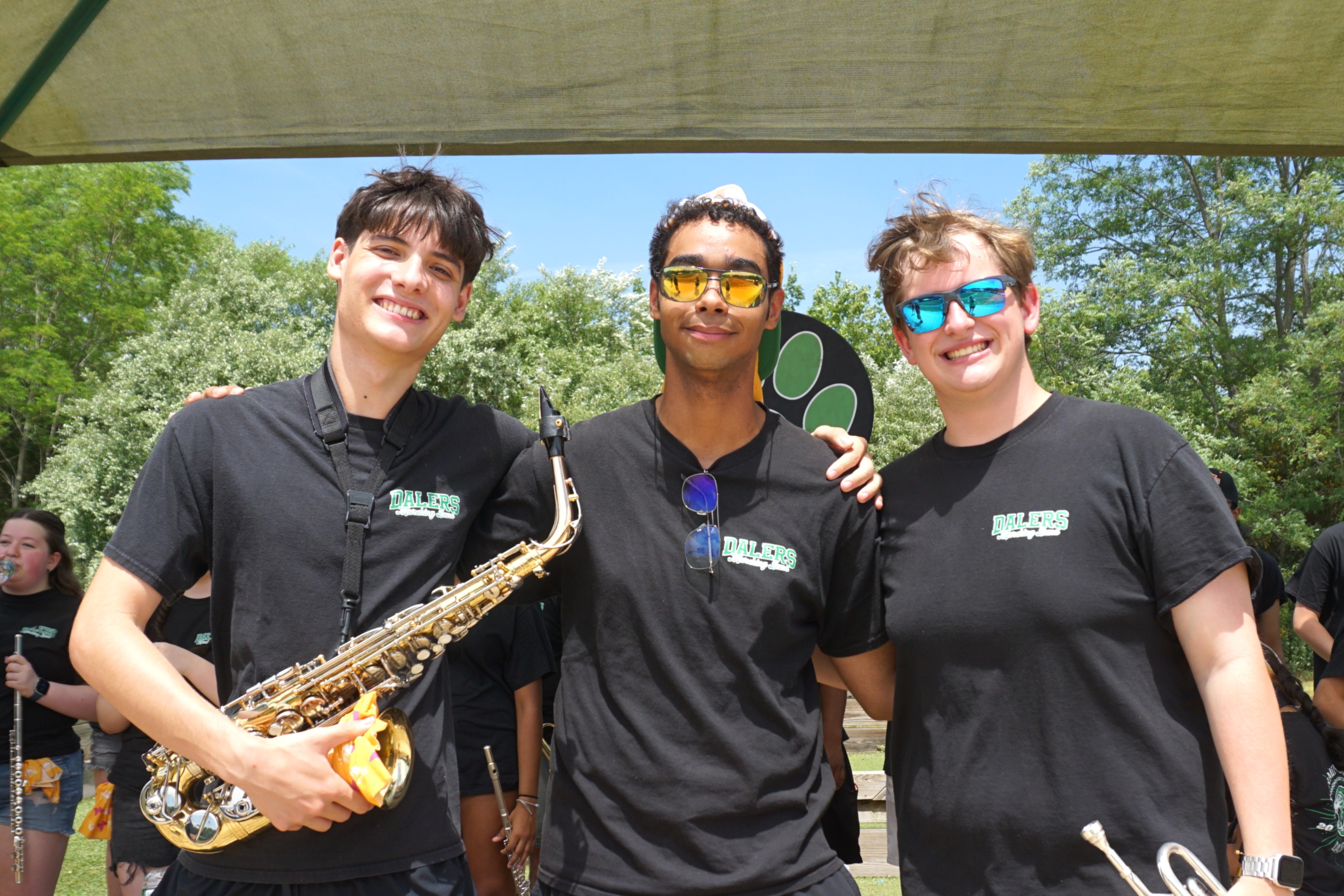 Three smiling young people in black t-shirts with 'DALERS' logo stand together outdoors, one holding a saxophone.