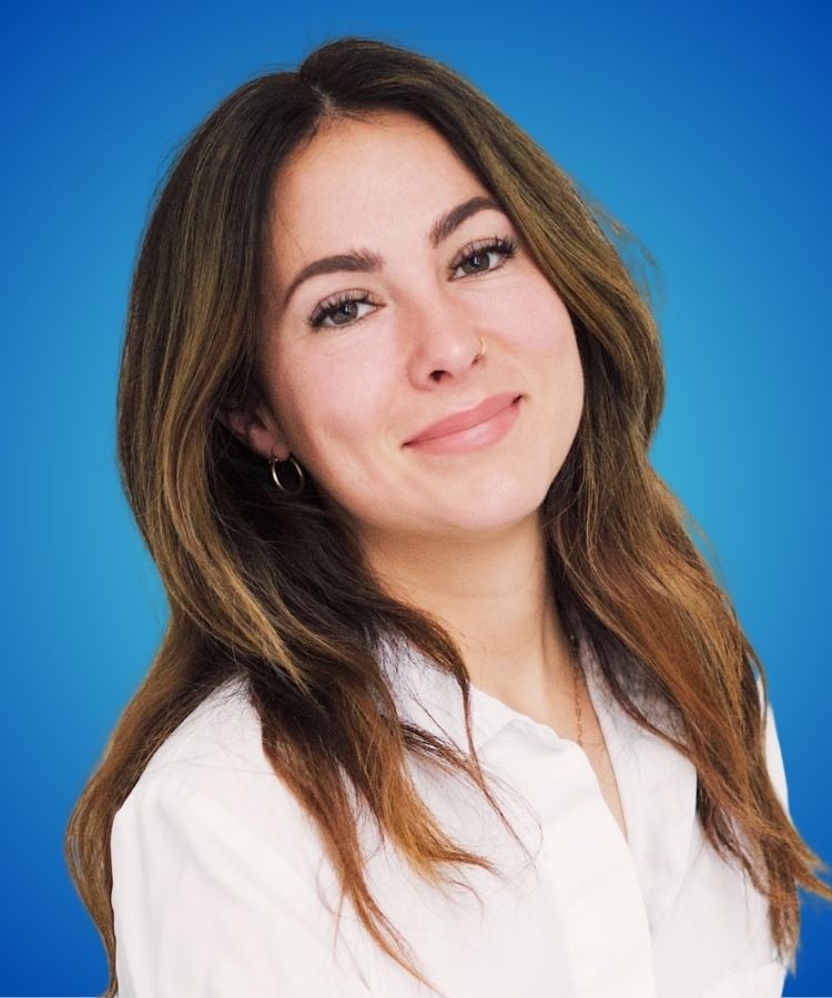 A close-up portrait of a smiling woman with long brown hair, wearing a white shirt against a bright blue background.