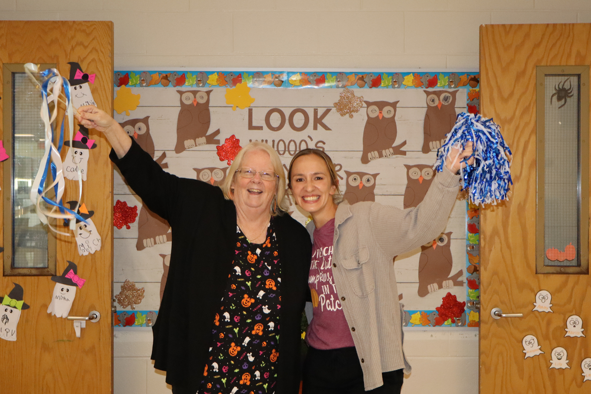 Two smiling women stand in front of an owl-themed bulletin board, holding celebratory items in a school hallway.