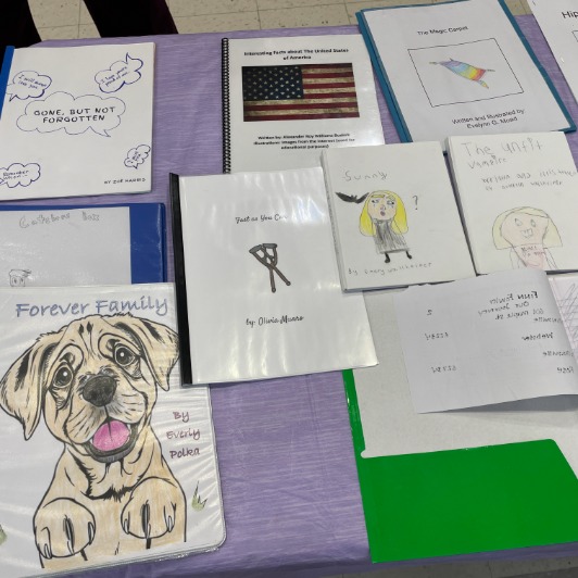 A collection of handmade books and projects are displayed on a table, featuring titles like 'Gone, But Not Forgotten' and 'Forever Family'.