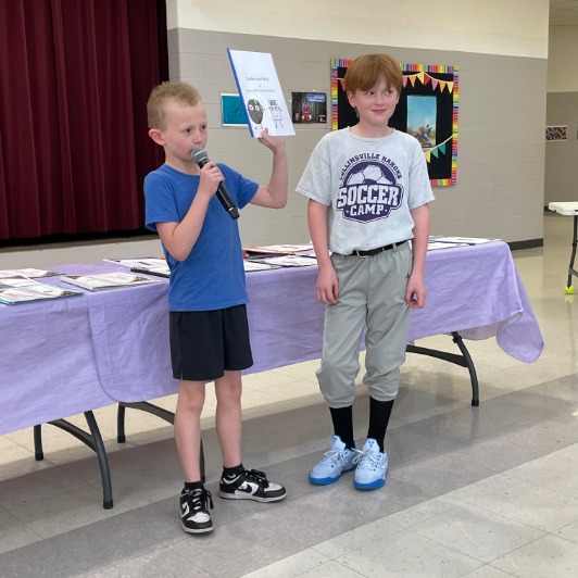 A young boy in a blue shirt holds a microphone and a book, presenting to an audience.