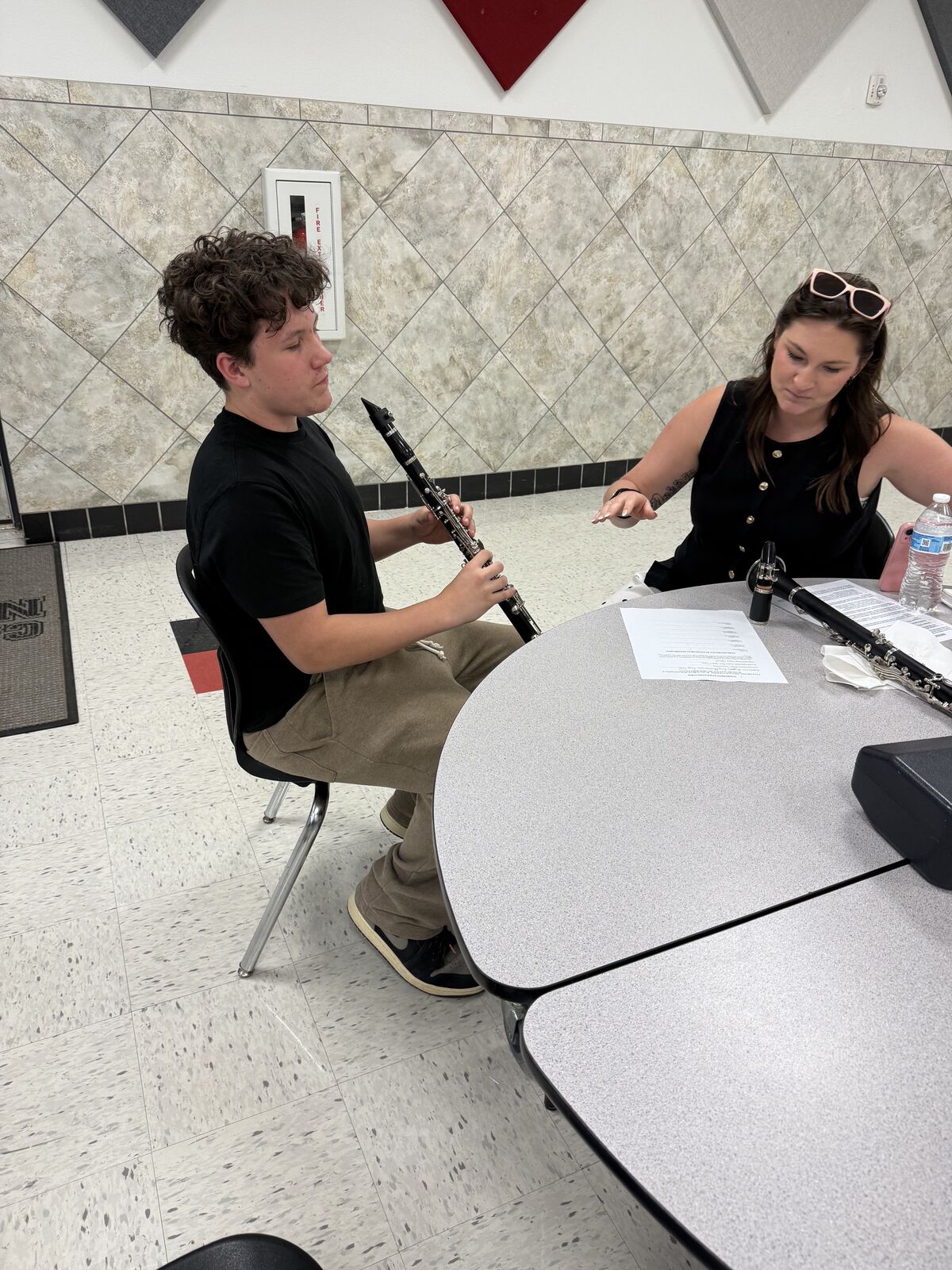 A music instructor guides a student playing the clarinet at a grey table in a brightly lit room.