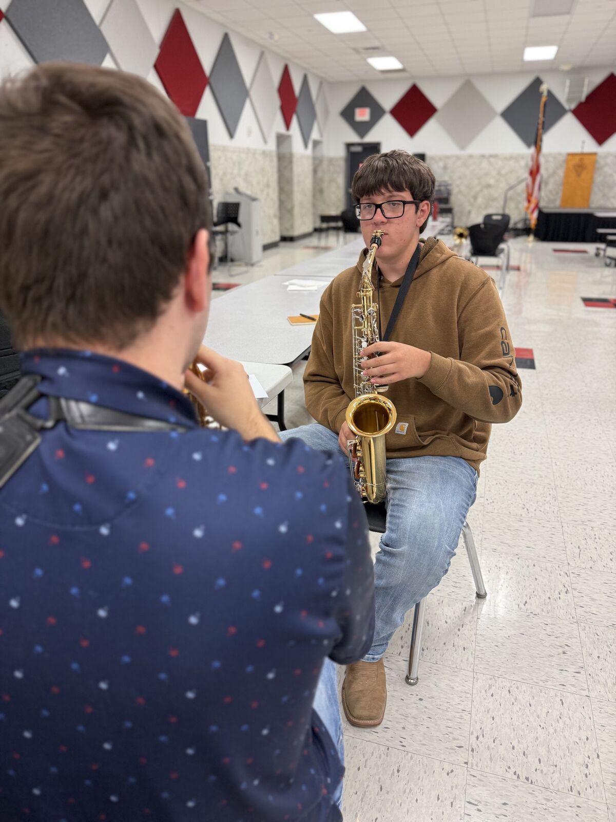 A young man in glasses and a brown hoodie plays the saxophone in a large room.