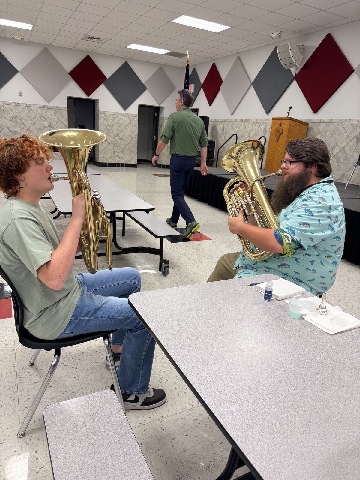 Two musicians in a room with patterned walls practice playing brass instruments at tables.