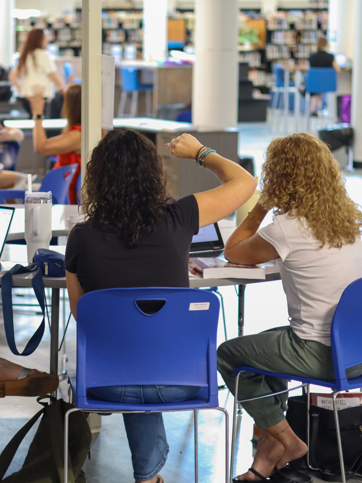 Two women sit at a table in a library, with one gesturing with her arm.