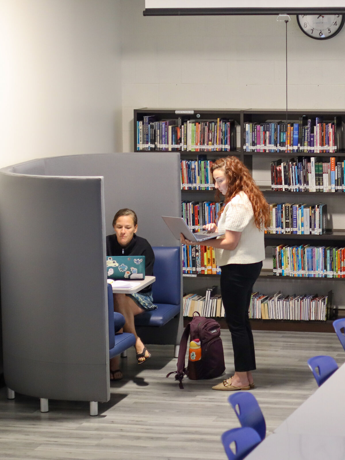 Two women work on laptops in a library study area with bookshelves in the background.