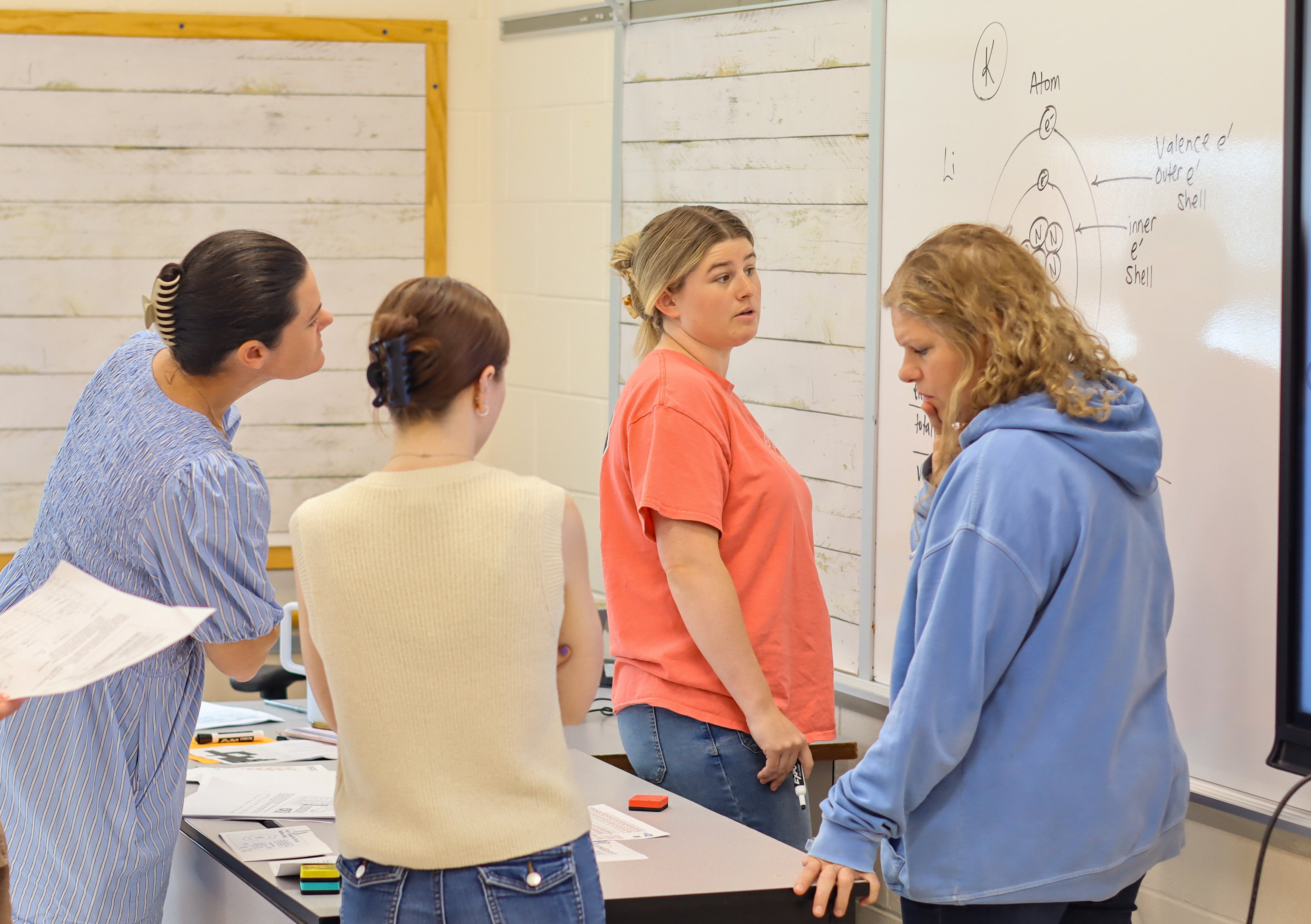 Four people gather around a table in a classroom, looking at a whiteboard with scientific diagrams.