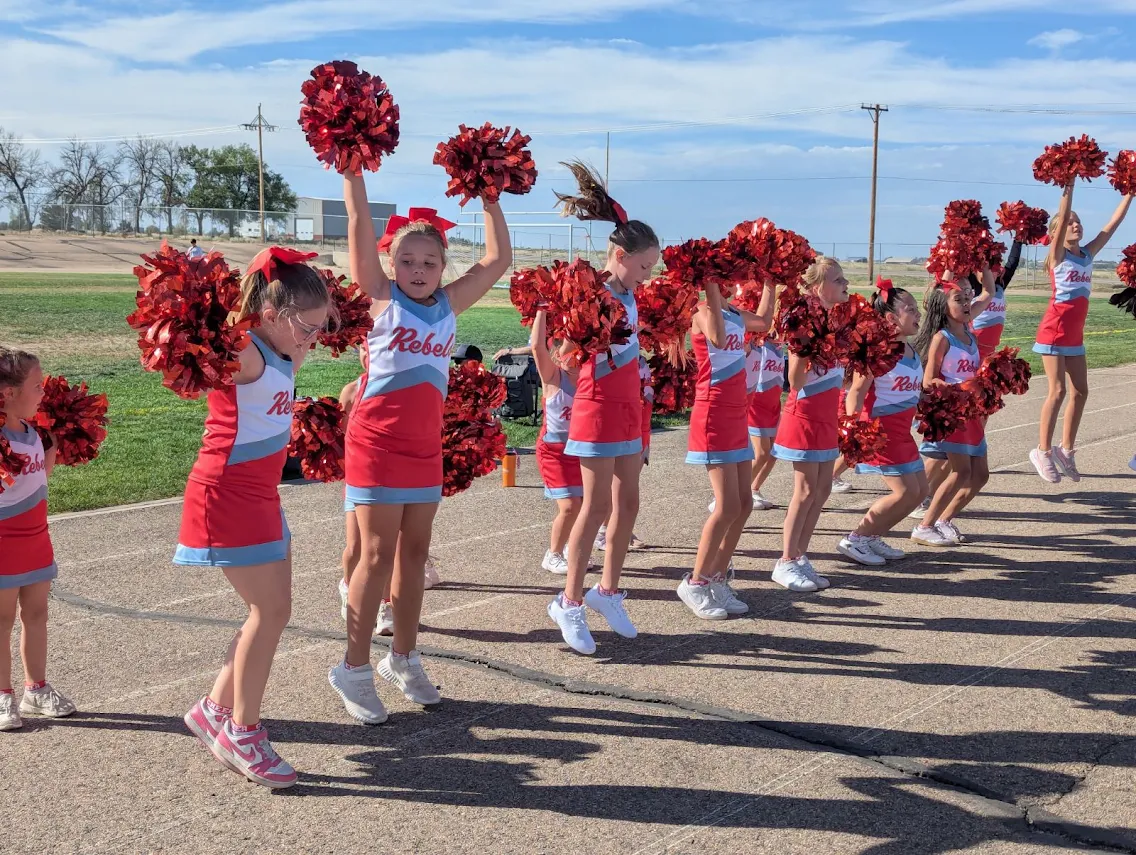 A line of young cheerleaders in red, white, and blue uniforms leap into the air outdoors, holding bright red pom-poms.
