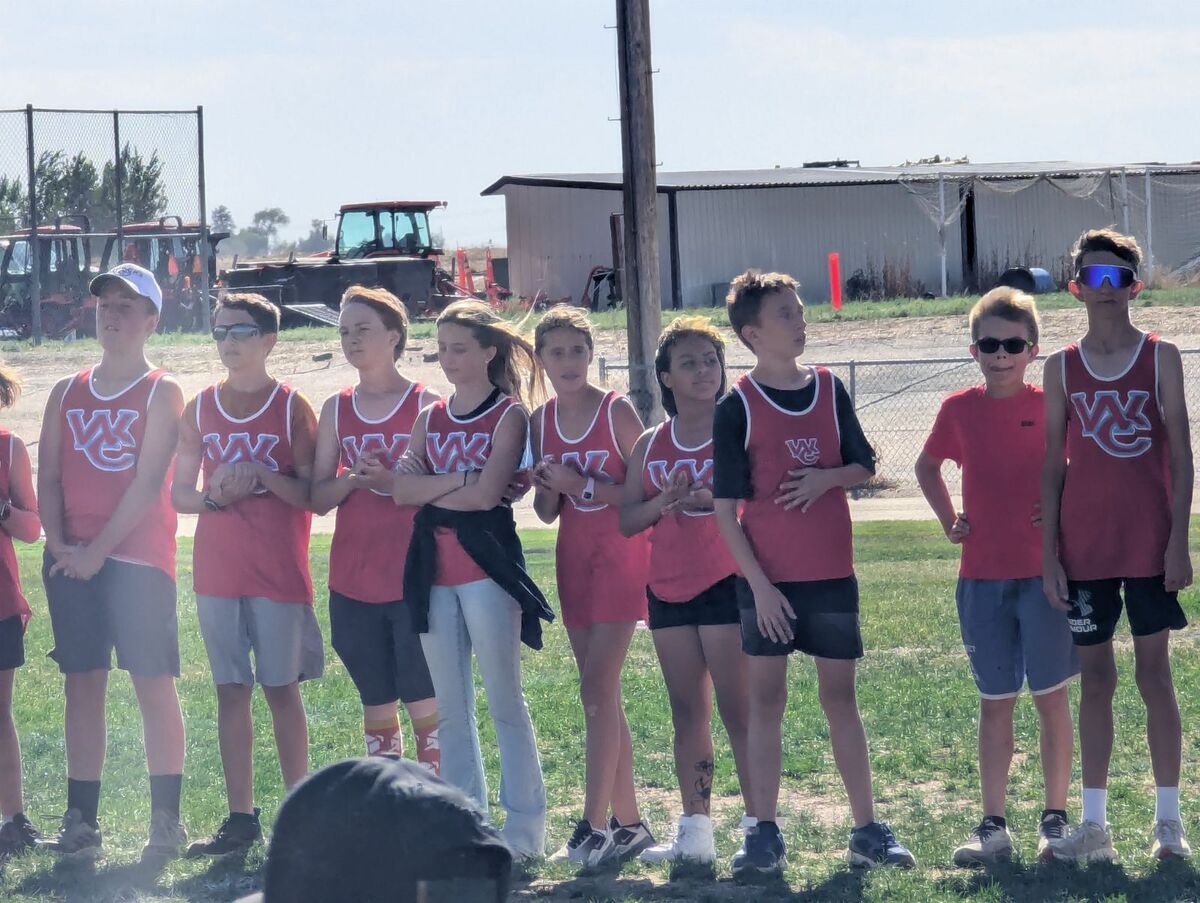 A line of young athletes in red and white athletic uniforms stands on a grassy field, watching an event.