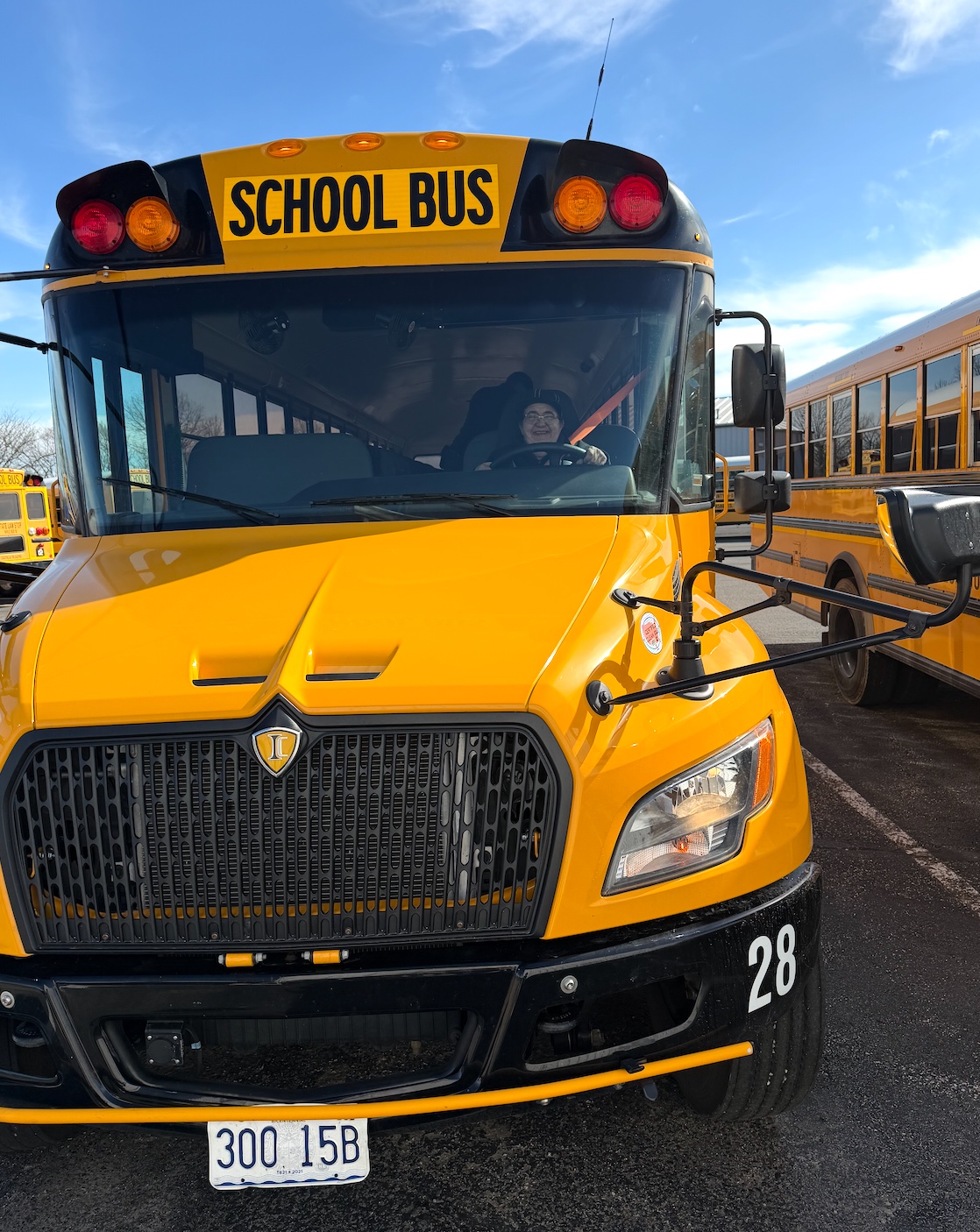 A person smiles while sitting in the driver's seat of a bright yellow school bus.