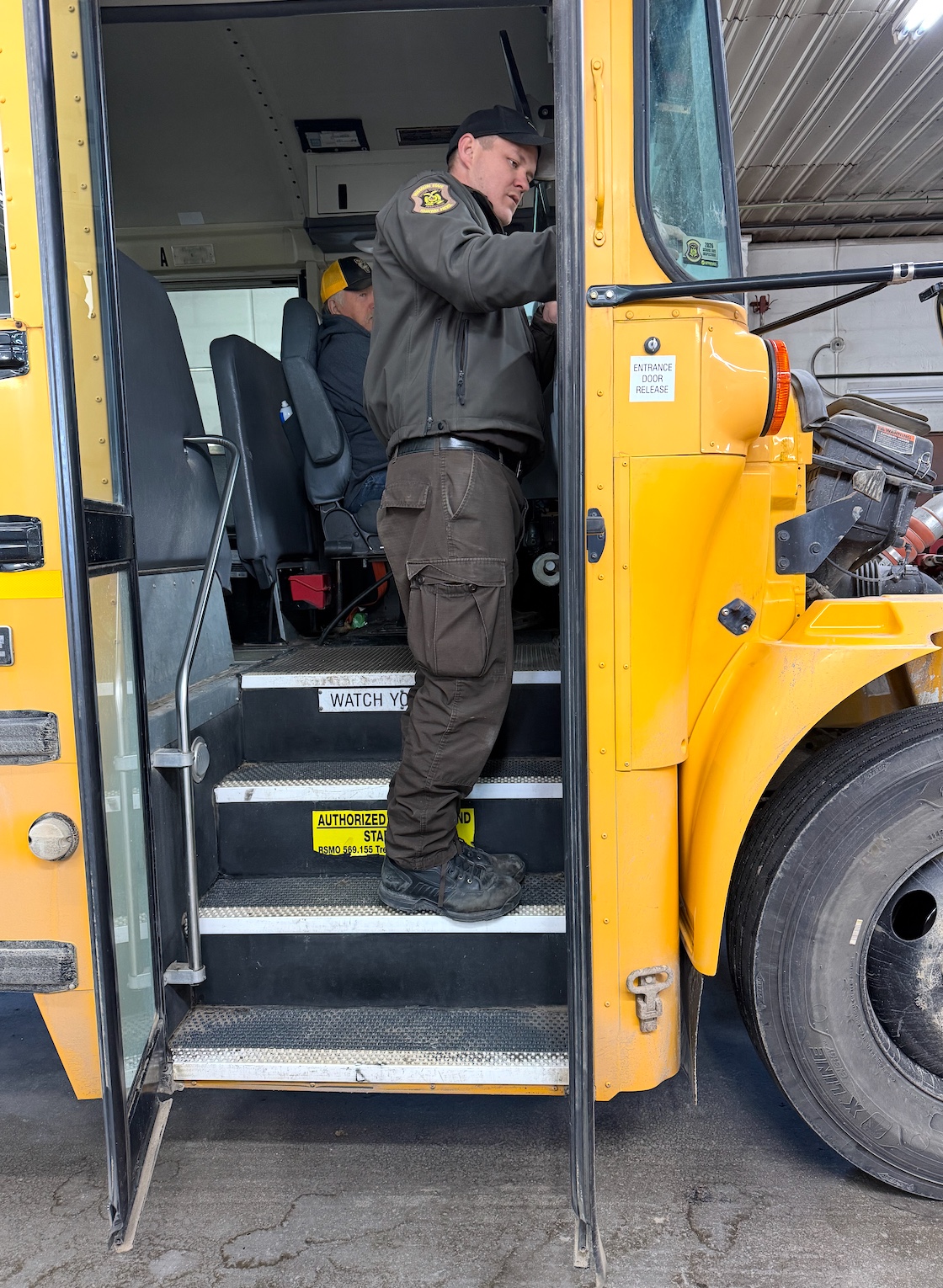 A person in a uniform stands on the steps of a yellow school bus, appearing to work on the bus.