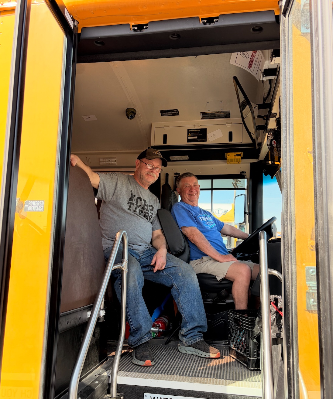 Two men smile while sitting inside the front of a yellow school bus.